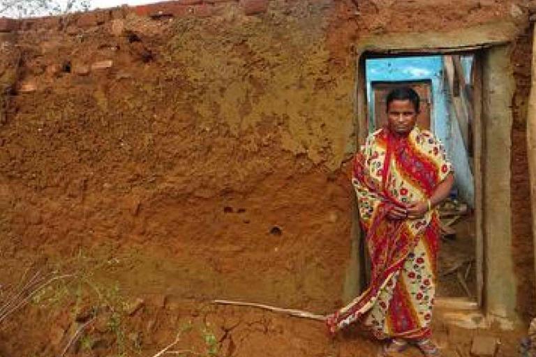 A woman in a patterned sari stands in the doorway of a partially collapsed mud brick house.