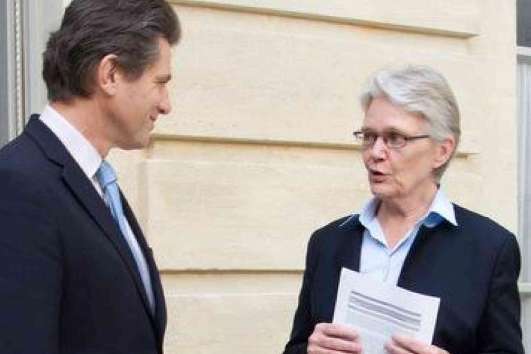 Two people in business attire stand outside a building, having a conversation; one holds printed documents.