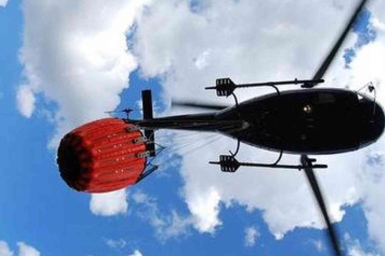 A helicopter carrying a large red water bucket flies overhead against a blue sky with scattered clouds.