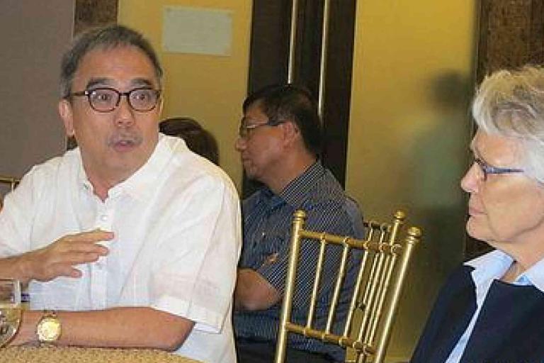 Two people are seated at a table, engaged in conversation during an indoor event. Other attendees are present in the background.