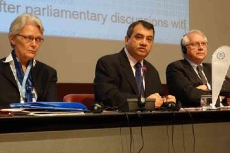 Three people in formal attire sit at a conference table with microphones, laptops, and documents, speaking at a meeting or panel discussion.