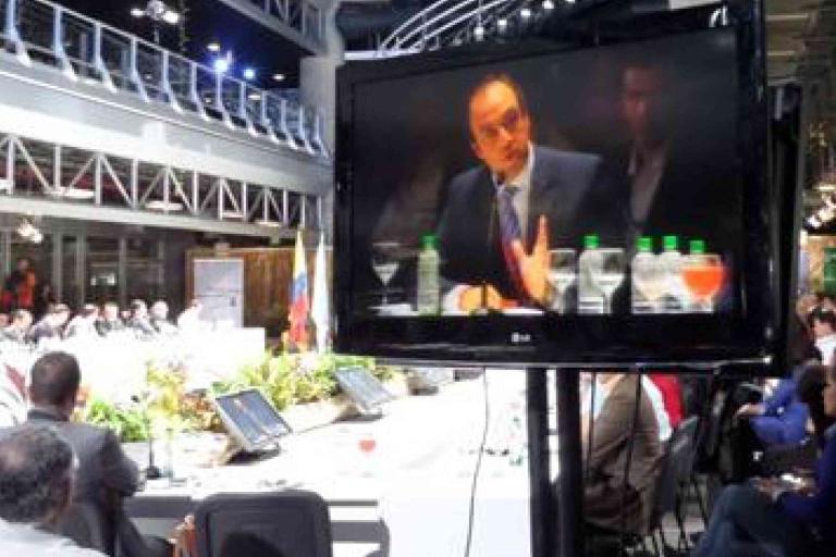 A large conference room with people seated in a meeting; a TV screen in the foreground displays a man in a suit speaking.
