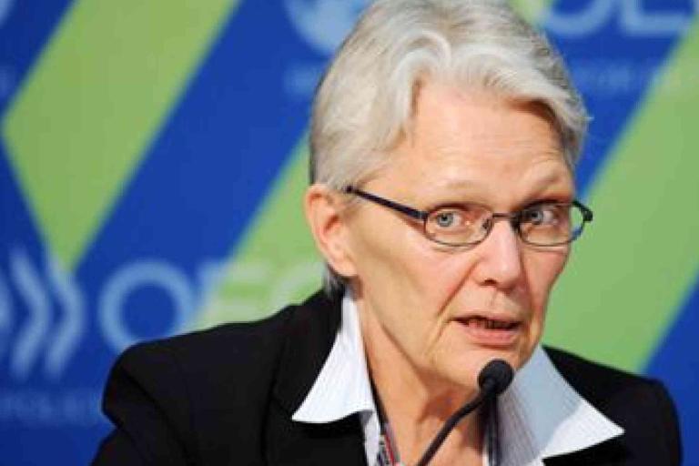 An older person with short gray hair and glasses speaks into a microphone, wearing a suit jacket and white collared shirt, with a blue and green background behind them.