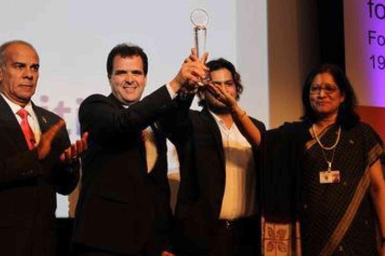 Four people in formal attire stand on stage; one man in the center holds up a trophy while others applaud during an award ceremony.