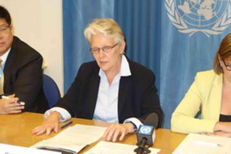 Three people sit at a table with documents and microphones in front of a UN flag, appearing to speak or present at a press conference.
