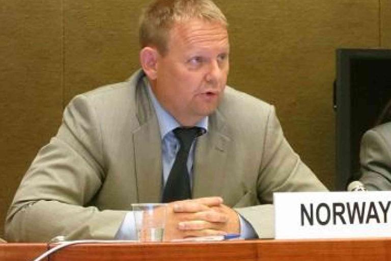 A man in a suit sits at a conference table with a nameplate labeled "NORWAY" in front of him, speaking into a microphone.