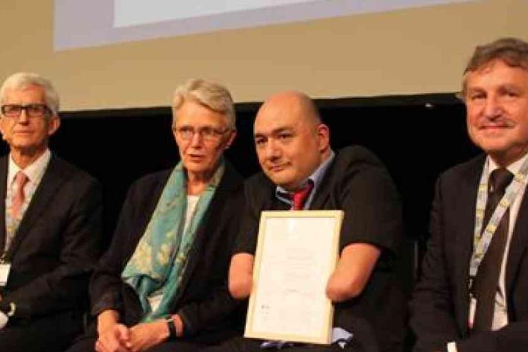 Four people sit together indoors; one person, holding a certificate, has no hands or forearms. All are dressed in formal attire and appear to be participating in an official event.