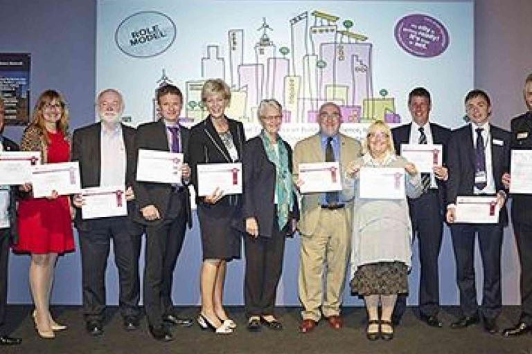 A group of eleven people stand in a row holding certificates, posing for a photo in front of a colorful illustrated backdrop at an event.