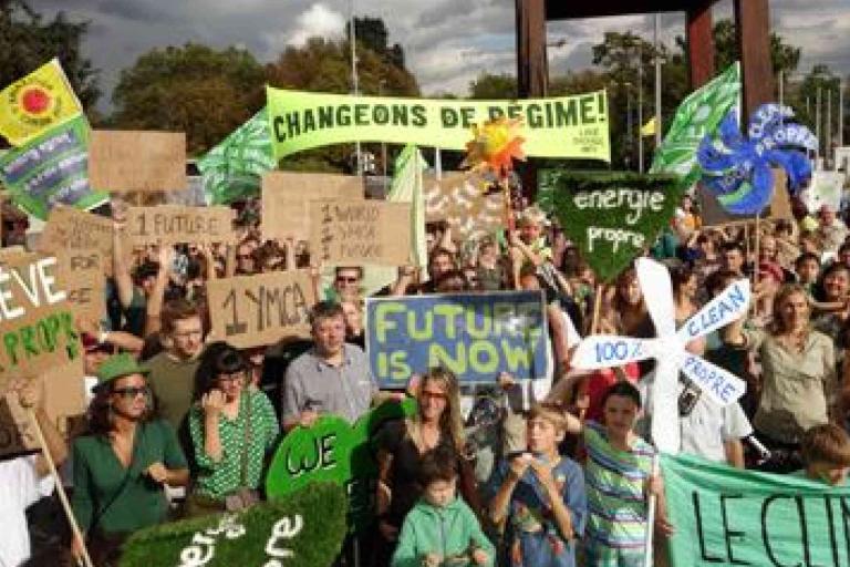 A large crowd holds signs demanding climate action and clean energy at an outdoor protest; banners include “FUTURE IS NOW” and “CHANGEONS DE RÉGIME!”.