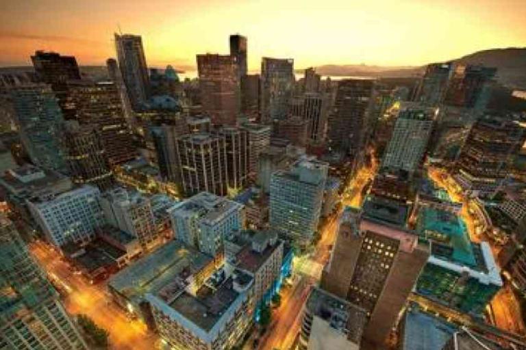 Aerial view of a city skyline at sunset, with tall buildings, illuminated streets, and a body of water in the background.