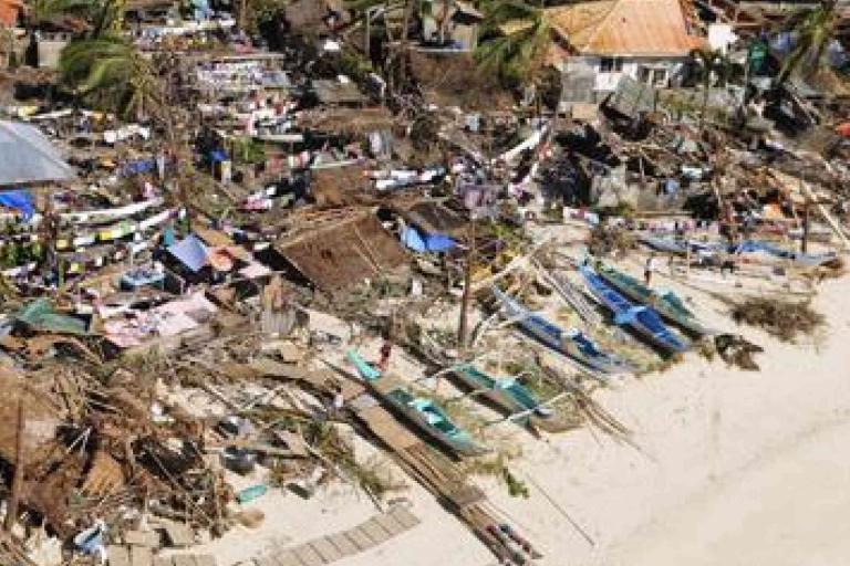 Aerial view of a coastal area with damaged houses, scattered debris, and boats on a sandy beach, likely after a natural disaster.