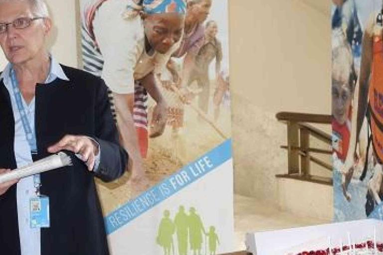 A person stands indoors holding a folder, with banners featuring people and the text "RESILIENCY IS FOR LIFE" displayed in the background.