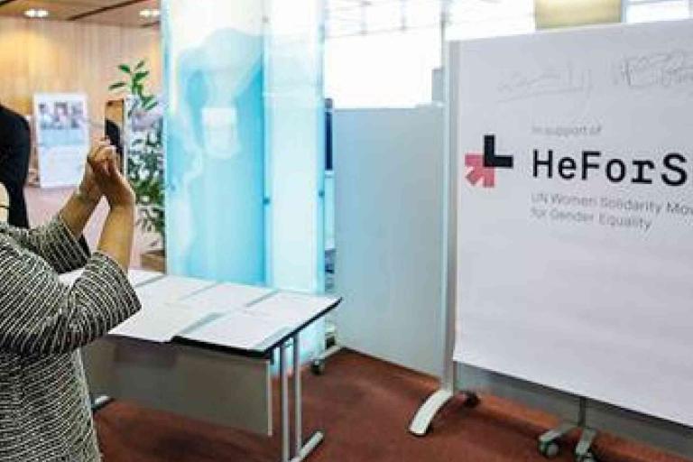 A person takes a photo of a large "HeForShe" sign at an indoor event promoting UN Women's solidarity movement for gender equality.