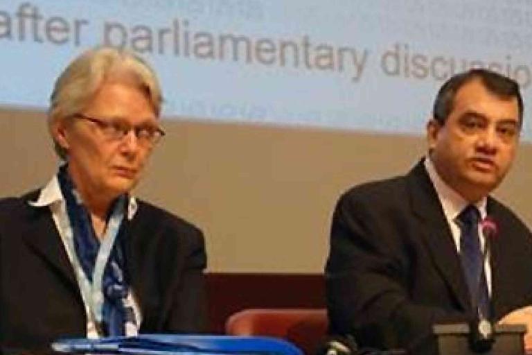 Two people in business attire are seated at a conference table, speaking in front of a projection screen with partial text visible in the background.