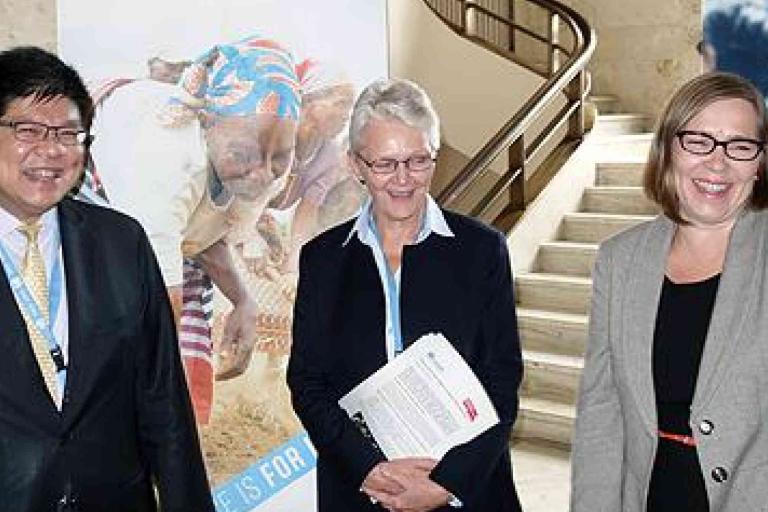 Three adults in business attire stand indoors, smiling and conversing, with a staircase and a large poster featuring children in the background.