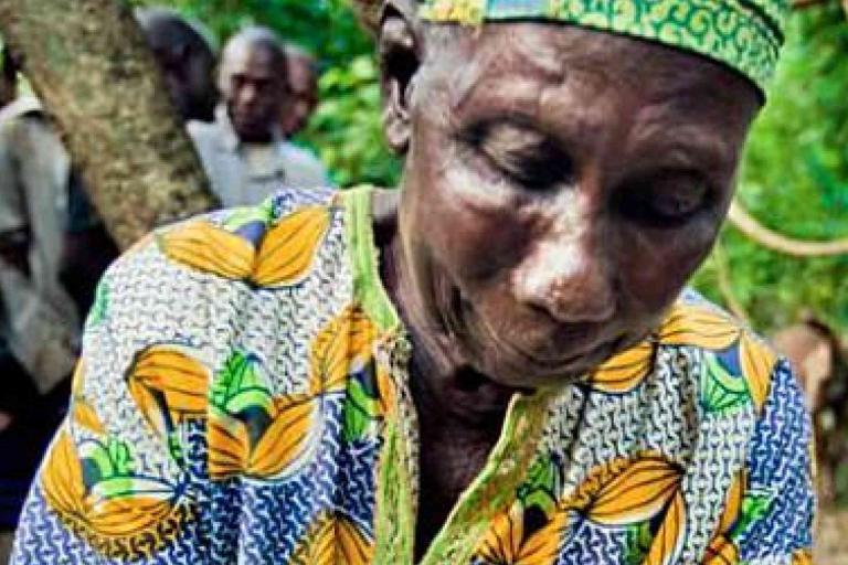 An older person wearing a patterned shirt and headscarf looks down while outdoors, with several people and trees visible in the background.