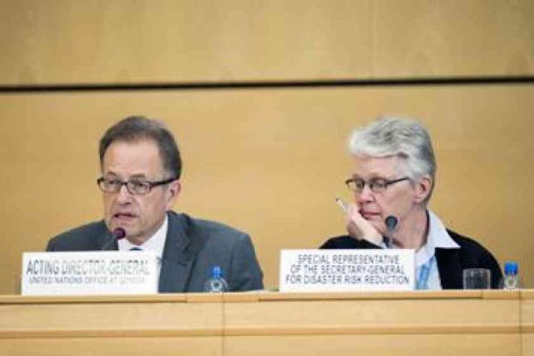 Two officials sit at a conference table with nameplates reading "Acting Director-General, United Nations Office at Geneva" and "Special Representative for Disaster Risk Reduction.