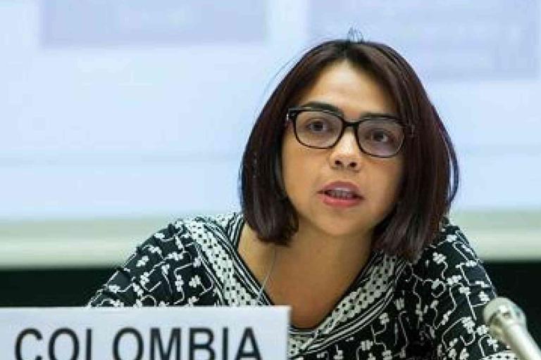 A woman wearing glasses speaks into a microphone at a conference, seated behind a sign that reads "COLOMBIA.
