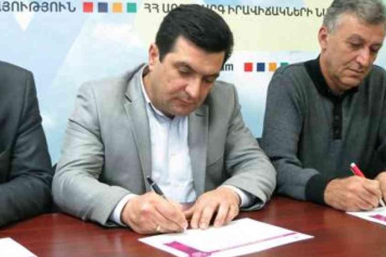 Three men sit at a table, signing documents with pens. A colorful banner with Armenian text is visible in the background.