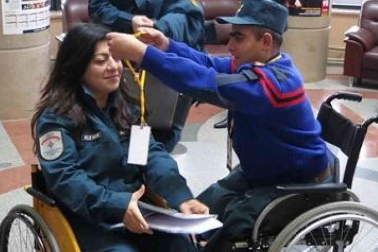 A man in a wheelchair places a lanyard around a woman's neck; she is also in a wheelchair. They appear to be in a public indoor space.