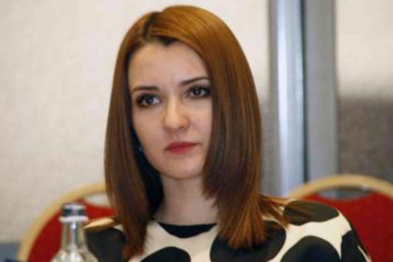 A woman with straight, shoulder-length brown hair sits in front of red chairs, wearing a black and white patterned top. A bottle is partially visible in the foreground.