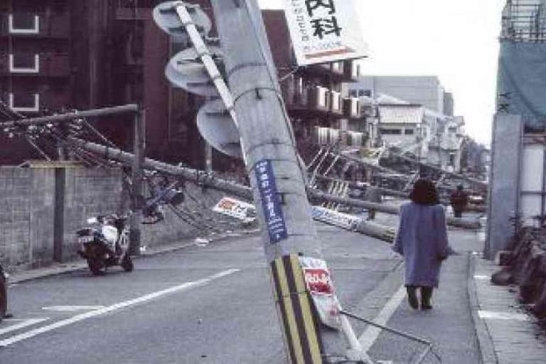 A street with multiple fallen utility poles leaning across the road; a person walks along the sidewalk beside the damage.