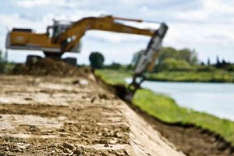 An excavator works on leveling or moving soil near a body of water, with a dirt embankment in the foreground and greenery in the background.