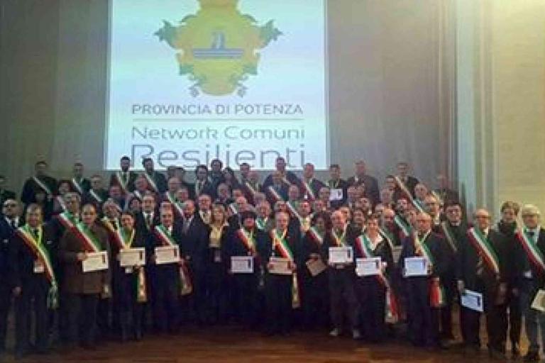 A large group of people poses on stage holding certificates in front of a screen displaying "Provincia di Potenza Network Comuni Resilienti.