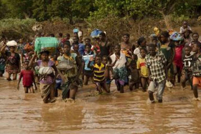 A large group of people wade through muddy floodwaters, carrying belongings on their heads and in their hands, surrounded by dense vegetation.
