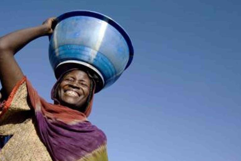 A smiling person wearing a colorful garment balances a large blue basin on their head against a clear blue sky.