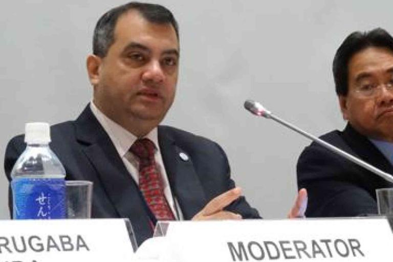 Two men in suits sit at a conference table with microphones; one is speaking and labeled as "MODERATOR" on the nameplate in front of him.