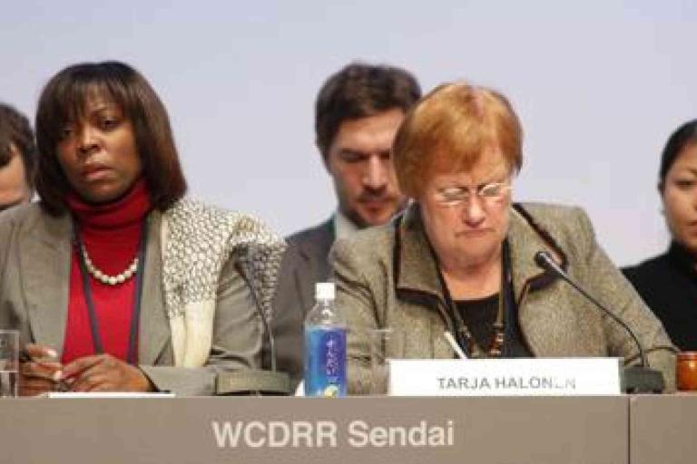 A group of people seated at a conference table with nameplates and water bottles; the nameplate in focus reads "TARJA HALONEN" and the backdrop says "WCDRR Sendai.