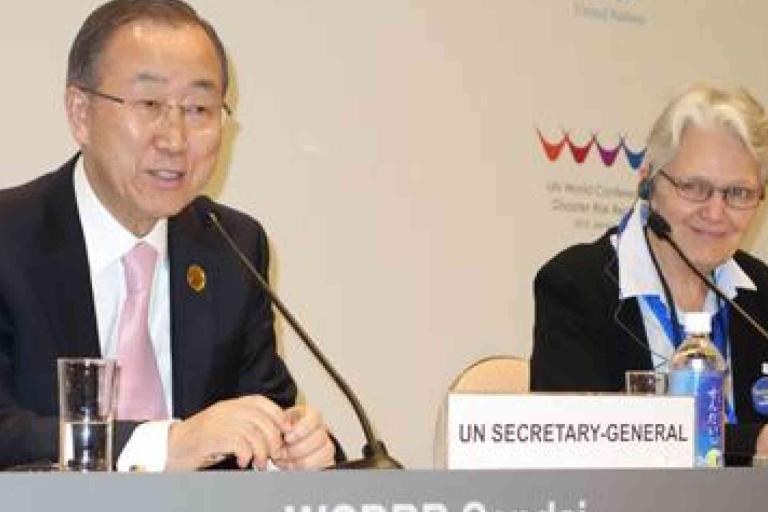 Two people sit at a conference table with microphones; the man on the left has a sign reading "UN SECRETARY-GENERAL" in front of him.