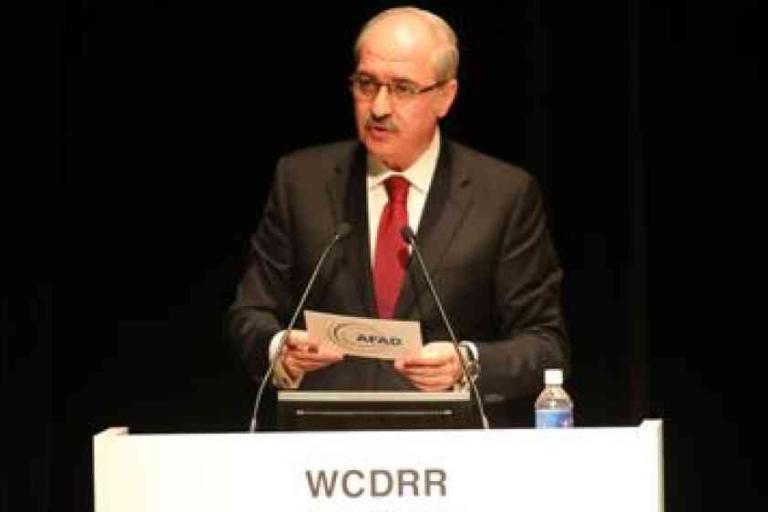 A man in a suit and tie speaks at a podium labeled "WCDRR," holding papers, with a microphone and a water bottle on the podium.