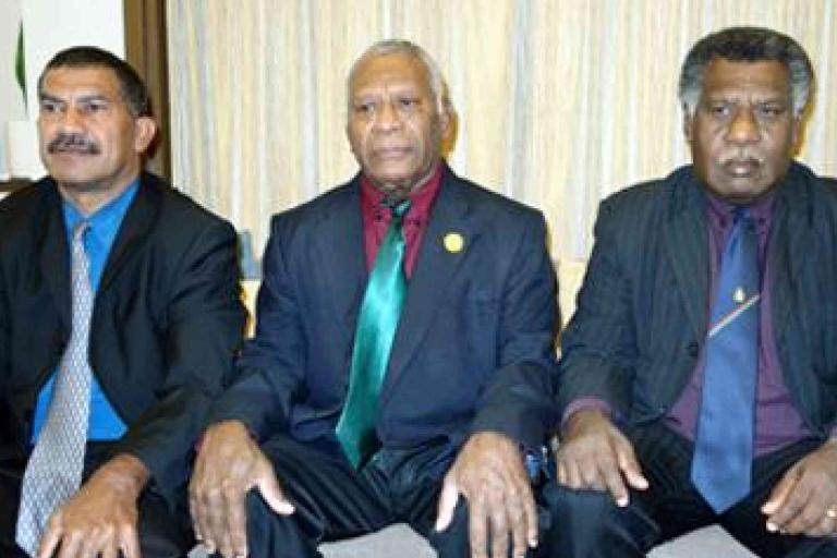 Three men in suits sit side by side on a couch, facing the camera with neutral expressions. The background features beige curtains.