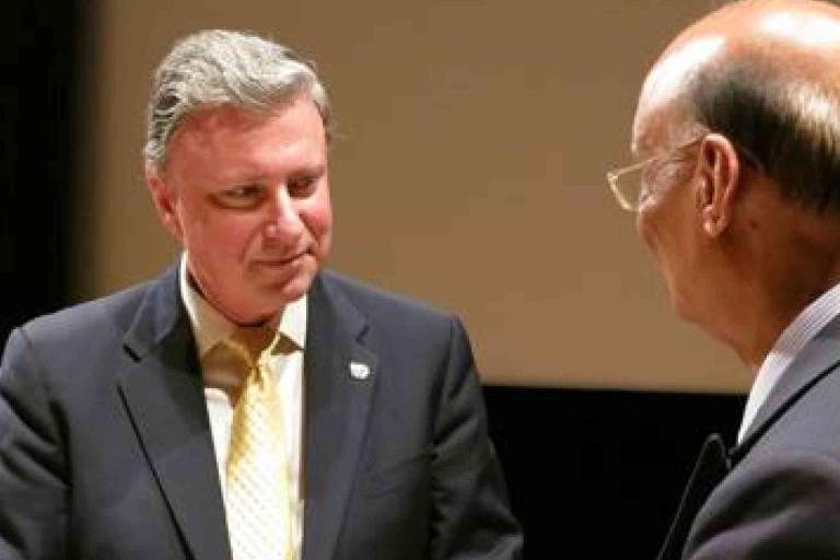 Two men in suits are engaged in conversation; one faces the camera while the other is seen from the side.