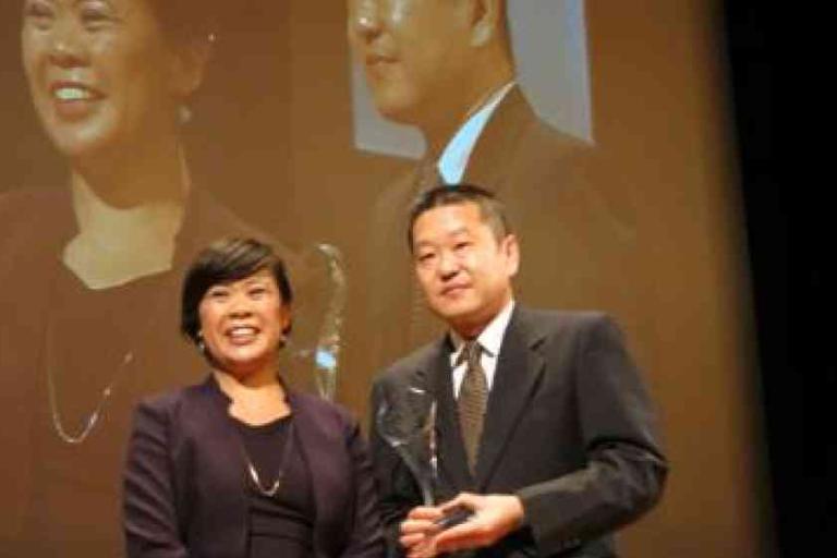 Two people in formal attire stand together on stage holding an award, with their image projected on a large screen behind them.