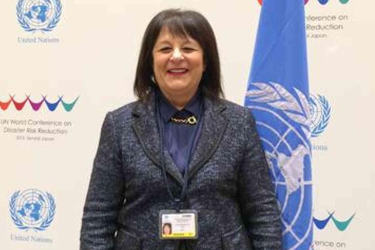 A woman in a dark suit stands in front of United Nations banners, smiling at the camera with an ID badge visible on her chest.