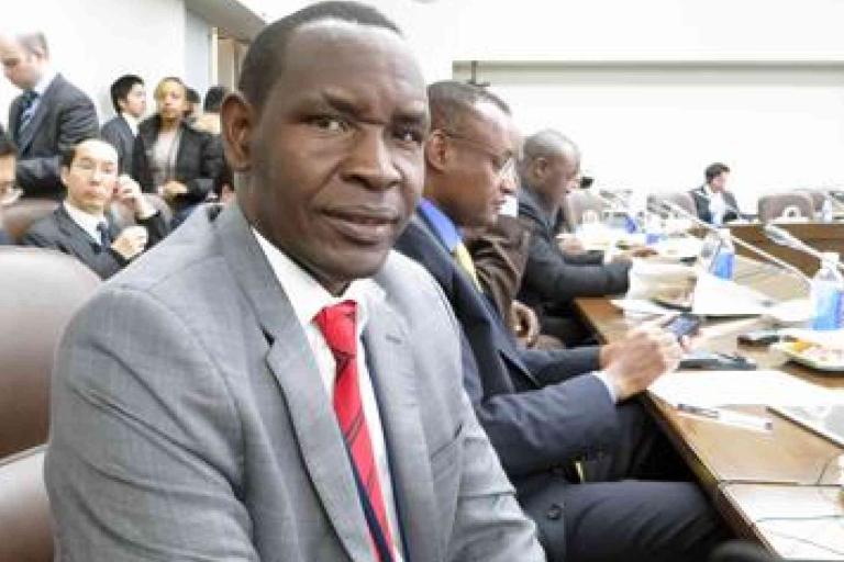 A man in a gray suit and red tie sits at a conference table with other attendees, some using laptops and tablets, in a formal meeting room.