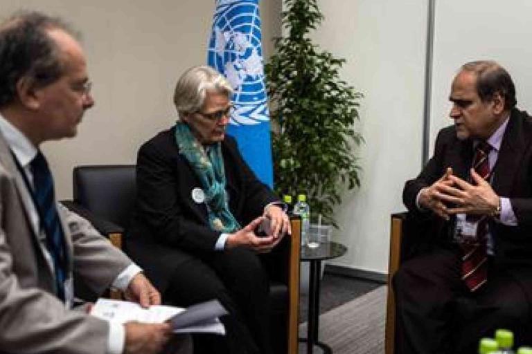 Three people in formal attire have a discussion in an office setting with a UN flag and water bottles visible in the background.