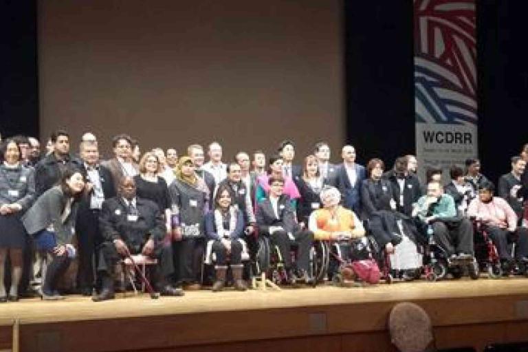 A large group of people, including several in wheelchairs, pose together on stage at an event with banners labeled "WCDRR" in the background.