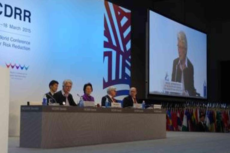 A panel of five people sit at a conference table on stage with a large screen displaying a speaker; multiple national flags are visible in the background.