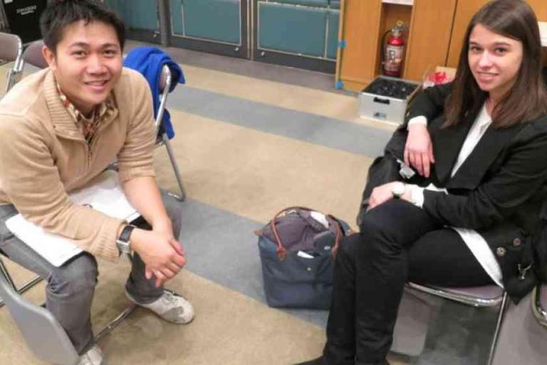 Two people sit on chairs indoors, facing the camera. Both have bags placed near them and appear to be in a casual, informal setting.