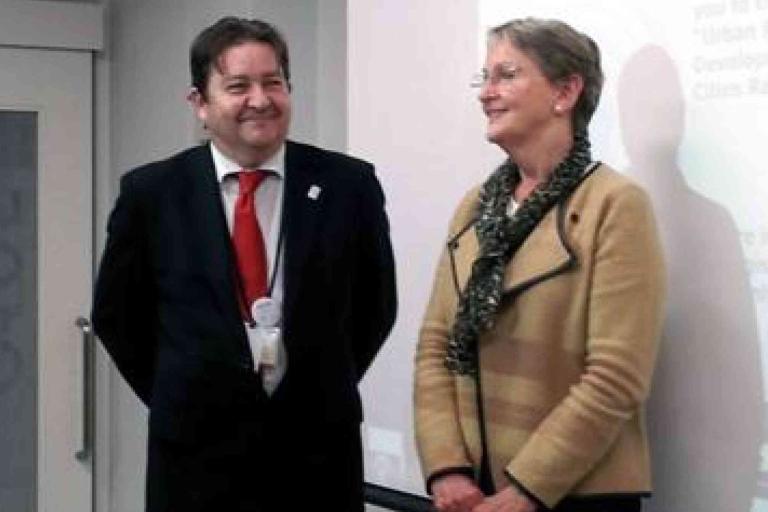 Two people stand indoors in front of a projected screen, both smiling. The man wears a suit and red tie; the woman wears glasses, a tan jacket, and a patterned scarf.