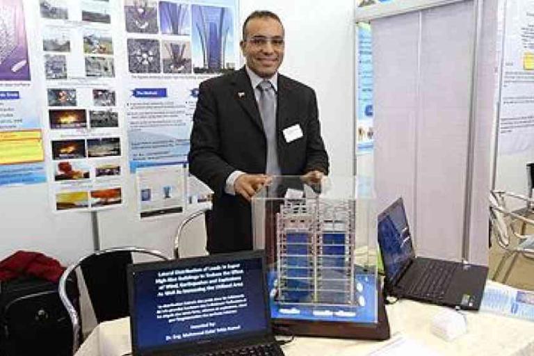 A man in a suit stands behind a table displaying a building model and two laptops, with scientific posters on the wall behind him.