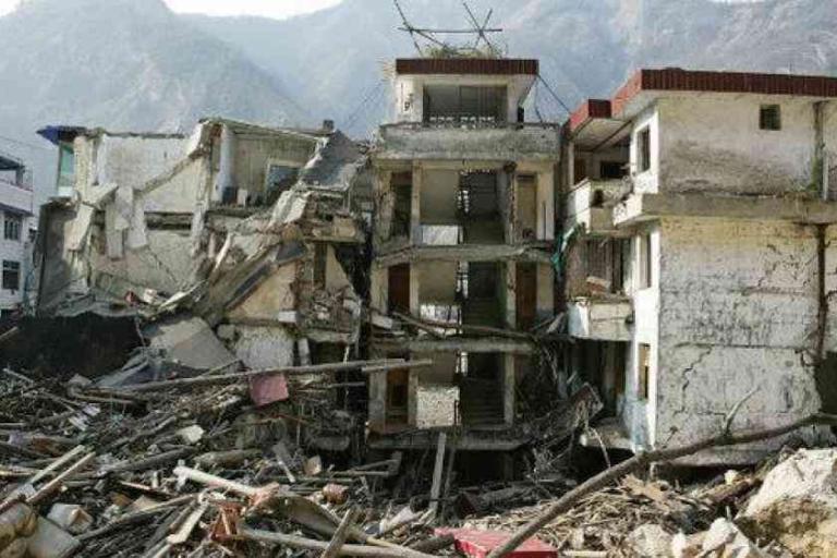 A partially collapsed building surrounded by debris and rubble, with mountains visible in the background.