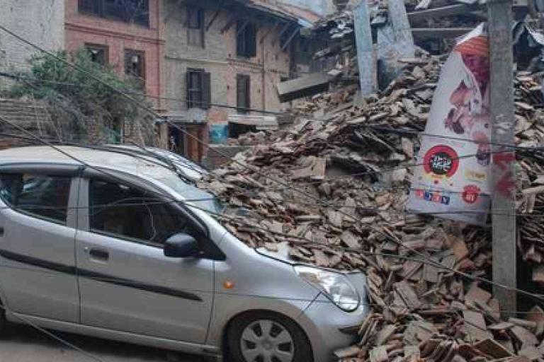 A silver car is parked beside a large pile of rubble from a collapsed building, with fallen debris and power lines surrounding the area.