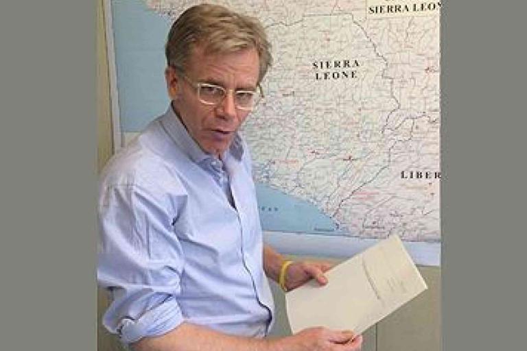 A person in a light blue shirt holding papers stands in front of a map showing Sierra Leone and surrounding regions.