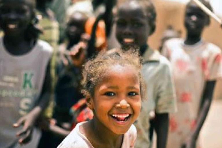 A young girl smiles at the camera while a group of children stand and interact in the background. The scene appears to be outdoors in a sunny setting.