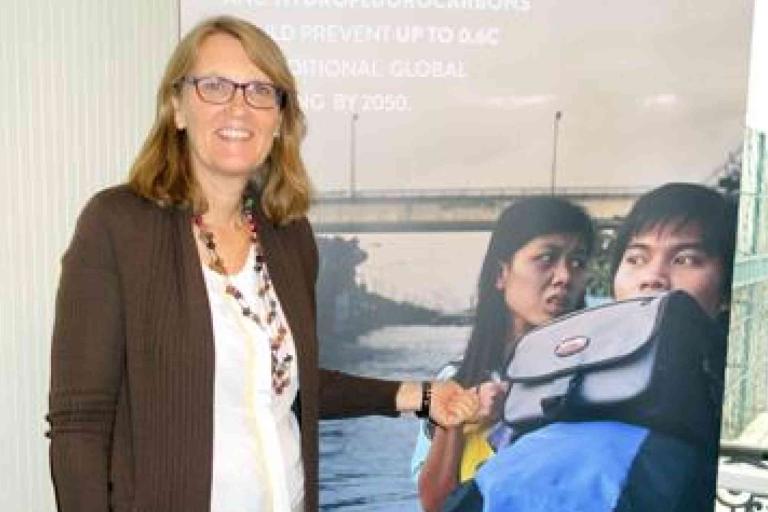 A woman standing indoors smiles and points at a poster featuring two people and an urban background.